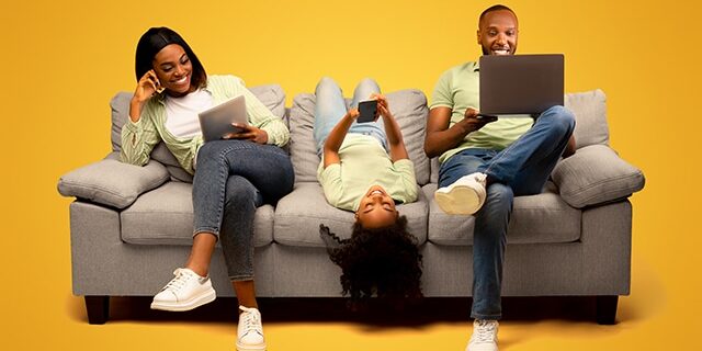 Gadgets addiction. Young black family of three using different electronic devices, sitting on sofa on yellow background Gadgets addiction. Young black family of three holding and using different electronic devices while sitting on sofa on yellow background. Parents and their daughter with modern gadgets