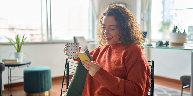 Young smiling pretty woman holding phone drinking coffee relaxing at home. Young smiling pretty woman holding smartphone technology drinking tea or coffee relaxing at home. Happy positive lady enjoying using mobile cell phone having fun while buying in ecommerce shop.
